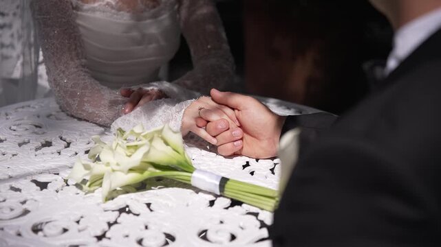 Close-up of a bride and groom holding hands next to a bridal bouquet of white calla lilies, showing an engagement ring.