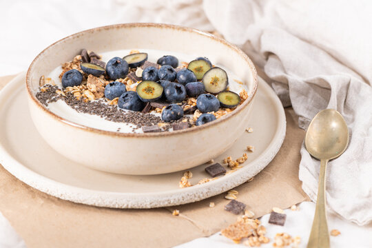 A nutritious breakfast yogurt bowl filled with fresh blueberries, crunchy granola, and chia seeds served in a rustic ceramic dish on a light textured fabric background for a healthy morning meal