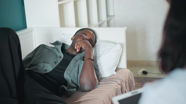 Black man patient receiving nurse consultation at bedside, blurred nurse in foreground holding tablet, patient reclining on bed with hand near mouth, chart review and verbal assessment, soft daylight,