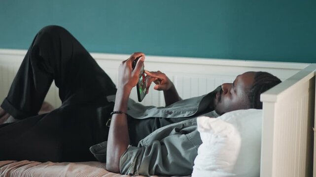 Black man lying bed scrolling phone, relaxed patient in home recovery, teal wall and white pillow, casual shirt and bracelet, calm indoor atmosphere, digital browsing and quiet leisure, awaiting