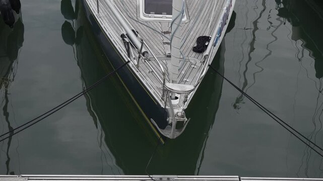 High angle view of a luxury sailing yacht bow moored in a calm harbor. Teak deck of a modern sailboat floating on still water with reflections, maritime lifestyle concept.