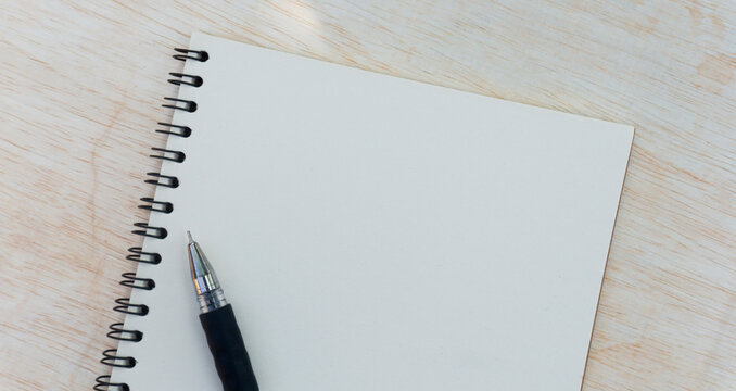 Close-up of a blank white spiral notebook with a black pen resting on a light wooden desk, representing note taking, planning, study, and office work.