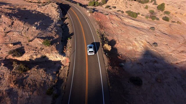 Aerial drone footage of Utah Scenic Byway 12 captures white car traveling along winding desert highway. Red rock formations and sparse vegetation line road as golden hour light illuminates landscape.