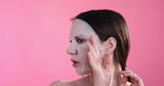 Young woman with sheet face mask on pink background