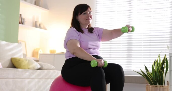 Overweight woman exercising with dumbbells on fitness ball indoors. Sports equipment