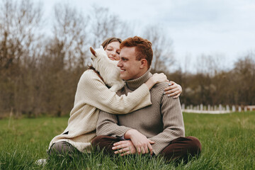 Obraz premium An affectionate couple sits on the grass in a sunny field, sharing a warm hug with a friendly goat kid between them. A candid lifestyle moment capturing connection, nature, and rural life.