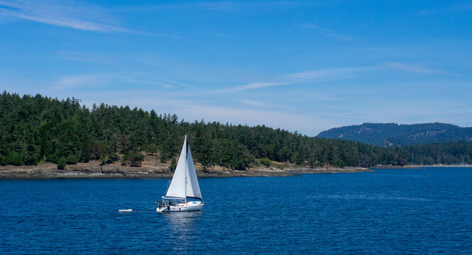 A sailboat under full sail, towing a dinghy, with paddleboard on the bow, is on a cruising vacation in the San Juan Islands, Washington State.