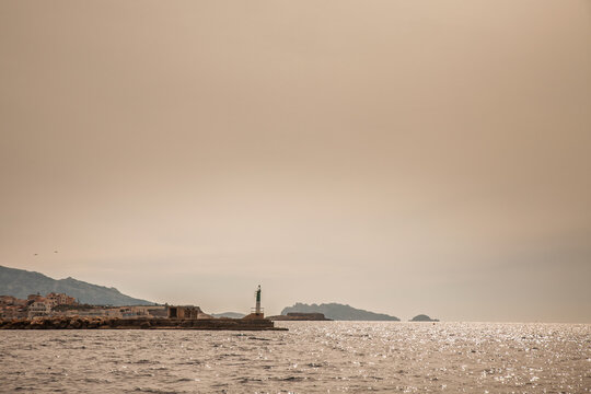 Atmospheric Mediterranean seascape with a green and white navigational beacon on a stone breakwater, coastal buildings and the rugged silhouettes of the Calanques in the distance in Marseille, France.