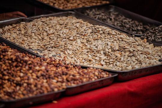 Roasted pumpkin seeds displayed for sale at a market stall, with trays of other seeds and nuts nearby. Pumpkin seeds are widely eaten as a snack and valued for protein, minerals and fiber.