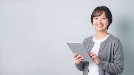 Obraz premium Portrait of a happy young Asian woman holding a digital tablet, looking at the camera with a bright smile. She is wearing casual clothes like a grey cardigan over a white t-shirt