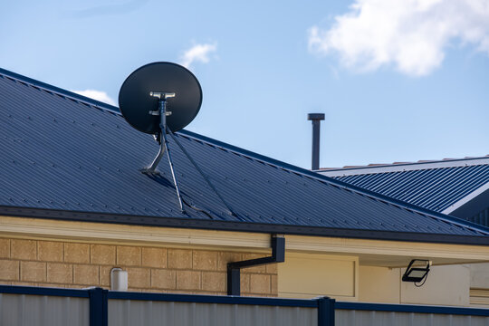 Satellite dish mounted on a corrugated metal roof of a suburban house, with a chimney flue and blue sky in the background, Melbourne, Australia