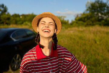 Obraz premium Young woman in a striped red shirt and straw hat smiles with eyes closed outdoors, standing in a sunny field. Natural light, lifestyle concept focused on happiness and leisure.