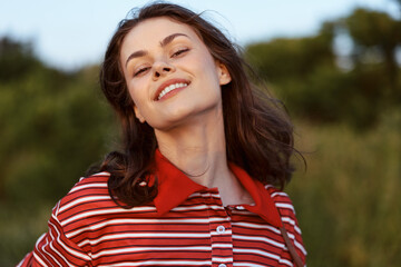 Obraz premium A smiling young woman in a striped polo shirt enjoys the warm sunlight outdoors. Lifestyle portrait of a happy girl in nature, concept of freedom, youth, joy and wellbeing.