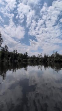 A beautiful, calm lake surrounded by coniferous trees against a backdrop of a blue sky with lovely clouds&mdash;the camera glides across the water's surface; shot from a watercraft. Ontario, Canada