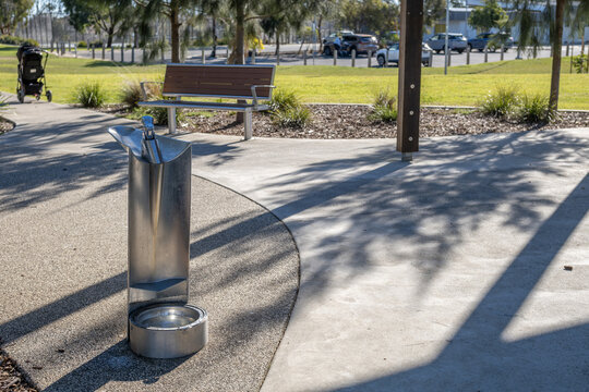 A stainless steel public drinking fountain with a integrated dog bowl base in a park, with park bench, grassy lawn&nbsp;in the background&nbsp;in a suburban park in Melbourne, Australia.