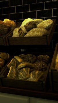Workers set out various types of bread in wooden baskets inside a bakery. The warm colors of the bread contrast with the dark tiles on the walls. Customers browse the fresh selection.
