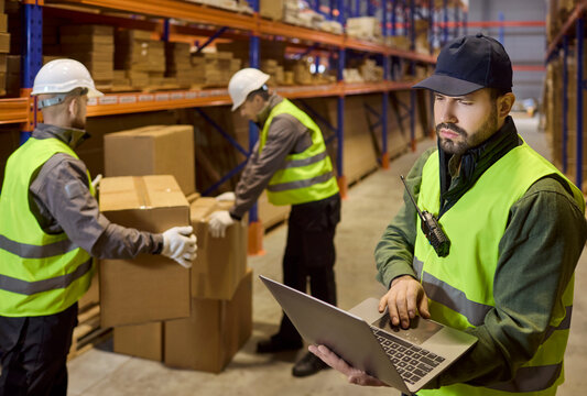 Warehouse manager reviews inventory on laptop. Workers move boxes on shelves, scan barcode labels with a scanner, update counts and coordinate logistics. Clear concept of warehouse efficiency.