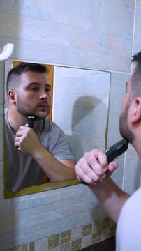 A young man trimming his beard with a trimmer in the bathroom in front of the mirror