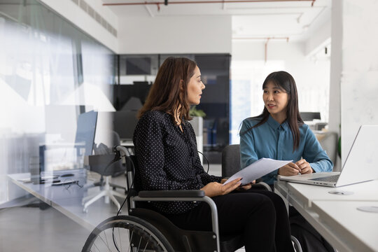 Young Arabic woman sitting in wheelchair, engaged in focused business discussion with Asian colleague at desk with laptop and documents. Workplace inclusivity, accessibility, equality and teamwork