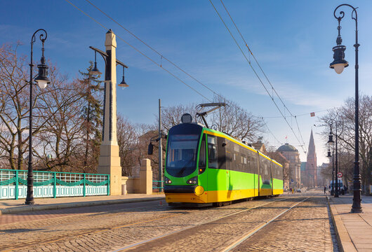 Green modern tram traveling across a historic bridge in central Poznan, Poland, with urban architecture in the background.