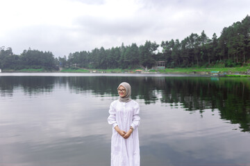 Fototapeta premium Portrait of a Muslim woman wearing a white hijab on the bank of a river. A young woman meets a summer daylight by the water.
