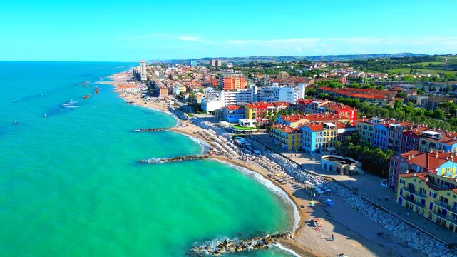 Delightful aerial push-in truck right pan left shot from Porto Recanati, flying above the Adriatic Sea and the shoreline washed by its waters, with stones, parasols, colored buildings, pleasant sky