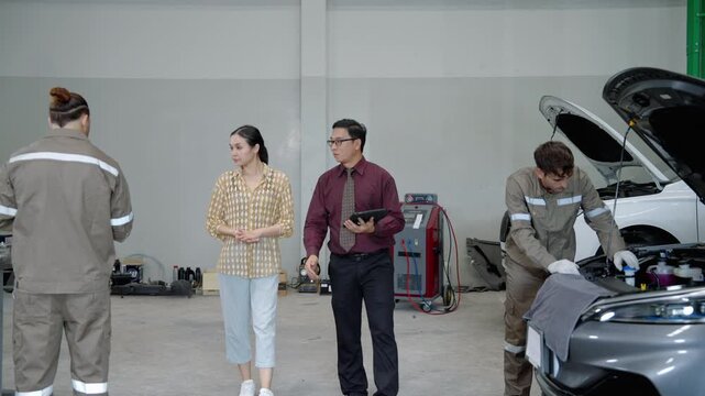 Automotive service advisor discusses vehicle maintenance with a customer while a technician works on a car in the background of a professional repair workshop.
