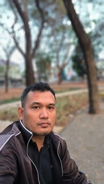 Portrait of a serious man sitting in a park during the daytime. Southeast Asian. 42 years old