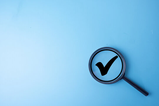 Top view of a magnifying glass focusing on a black check mark symbol on a solid light blue background with copy space. Concept of quality control, approval, verification, and successful choices.