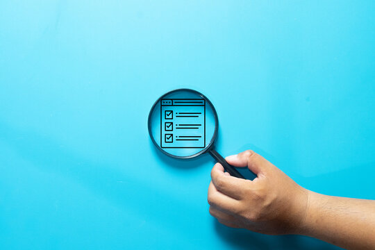 Hand holding a magnifying glass focusing on a digital checklist icon on a blue background. Concept of quality control, task management, online survey, and verification.