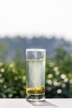 A transparent glass cup holds light green tea with tea leaves settled at the bottom, set against blurred outdoor greenery.