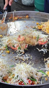 Asian street food vendor using spatulas to stir fry noodles and seafood on big hot pan.