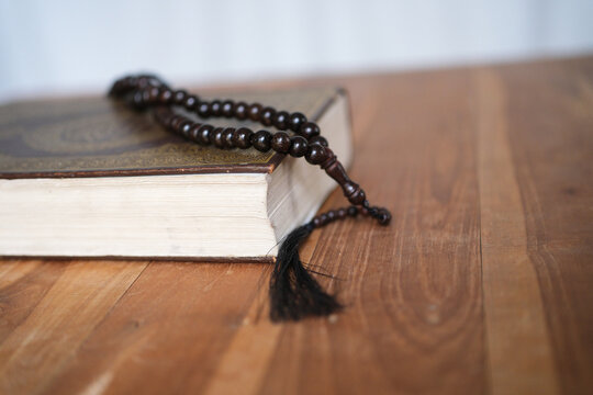 A prayer bead string rests on a closed Quran atop a wooden table; the focus is on the religious object in the foreground against a blurred background&mdash;a concept suitable for Ramadan, Hajj, and Islamic 
