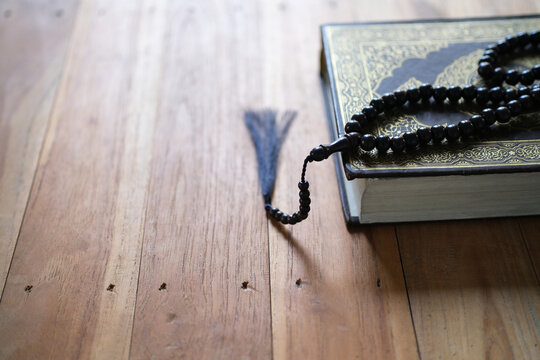 A prayer bead string rests on a closed Quran atop a wooden table; the focus is on the religious object in the foreground against a blurred background&mdash;a concept suitable for Ramadan, Hajj, and Islamic 