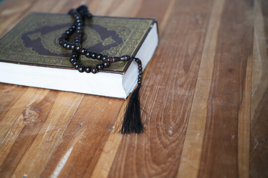 A prayer bead string rests on a closed Quran atop a wooden table; the focus is on the religious object in the foreground against a blurred background&mdash;a concept suitable for Ramadan, Hajj, and Islamic 