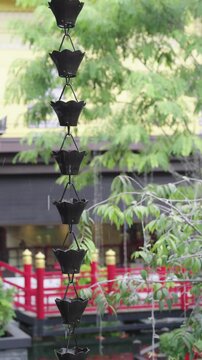 Decorative black metal rain chain hanging in a traditional Japanese garden during rainfall. Water drips through lotus cups with a blurred red bridge and green trees in the background.