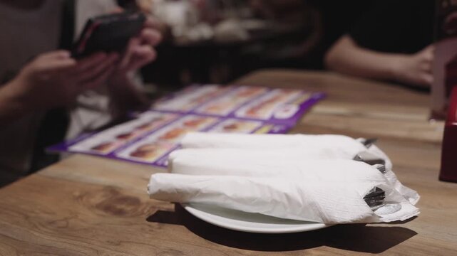 Silverware wrapped in white paper napkins on a plate at a restaurant table. In the background, a person looks at a menu and uses a smartphone. Casual dining and hospitality concept for websites.