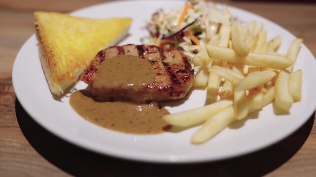 Grilled steak topped with savory black pepper sauce served on a white plate. Accompanied by crispy golden french fries, buttered toast, and fresh coleslaw salad. Professional food photography.