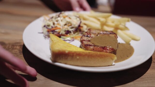 Grilled pork steak with gravy sauce served on a white plate with french fries, coleslaw salad, and toasted bread. A person holding the plate in a restaurant setting with warm lighting.