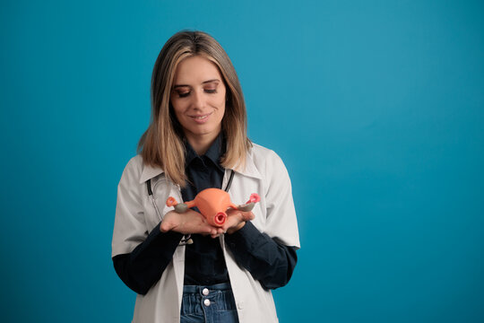 Female Doctor Wearing Lab Coat Holding Uterus Model on Blue Background. Smiling professional gynecologist showing anatomical replica of human uterus for medical education purpose