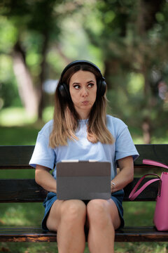 Thoughtful Young Woman With Headphones Working On Laptop Sitting On Park Bench. Pensive female student listening to music and using digital tablet with keyboard outdoors  