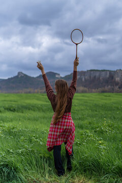 Triumphant woman holding badminton racket and shuttlecock aloft while standing in a lush green field under a cloudy sky