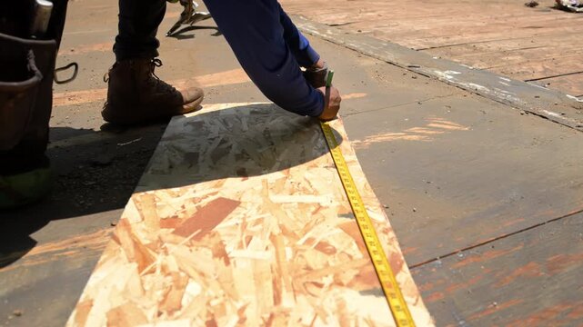 Construction worker installing roofing underlayment on a roof deck using OSB sheathing and measuring tape and a utility knife.