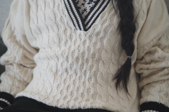 A cild sits indoors wearing a white sweater with a braided hairstyle