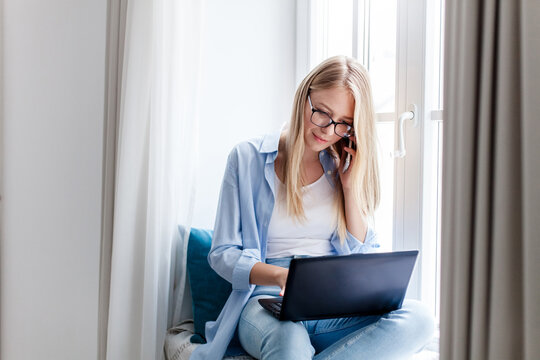 Freelancer working from home office. Young woman using laptop and talking on mobile phone. Cozy workplace on windowsill. Technology lifestyle. Concept of female small business, teleworking