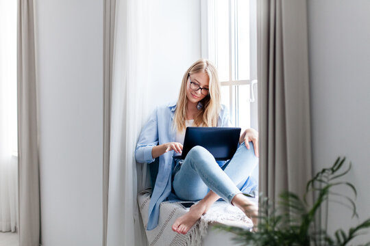 Young woman using laptop, working from home office. Relaxed freelancer. Workplace in living room on windowsill. Technology lifestyle. Female business, teleworking, online education, distance learning