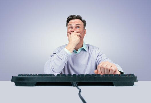 A frightened man sits at a computer, covering his mouth in shock and fear while using a keyboard. Concept of panic, anxiety, surprise, stress, and alarming news online. Shock caused by news.