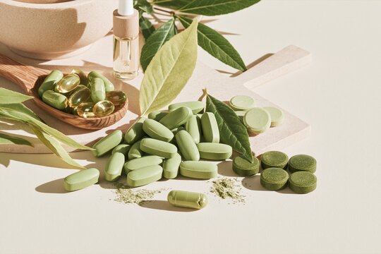 Assorted green supplement tablets and softgels scattered on a light surface with a wooden spoon and fresh leaves for herbal remedy marketing