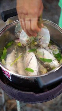 Cooking Thai spicy and sour short mackerel soup. Simmering lemongrass, kaffir lime leaves, and shallots, adding fresh mackerel, tomatoes, crushed chilies, spring onions, and holy basil in pot.