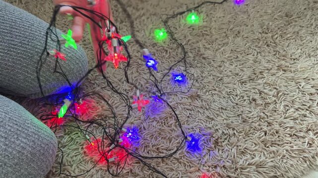 Woman untangling tangled colorful star shaped christmas lights on a beige carpet, preparing decorations for the holiday season at home in a cozy atmosphere